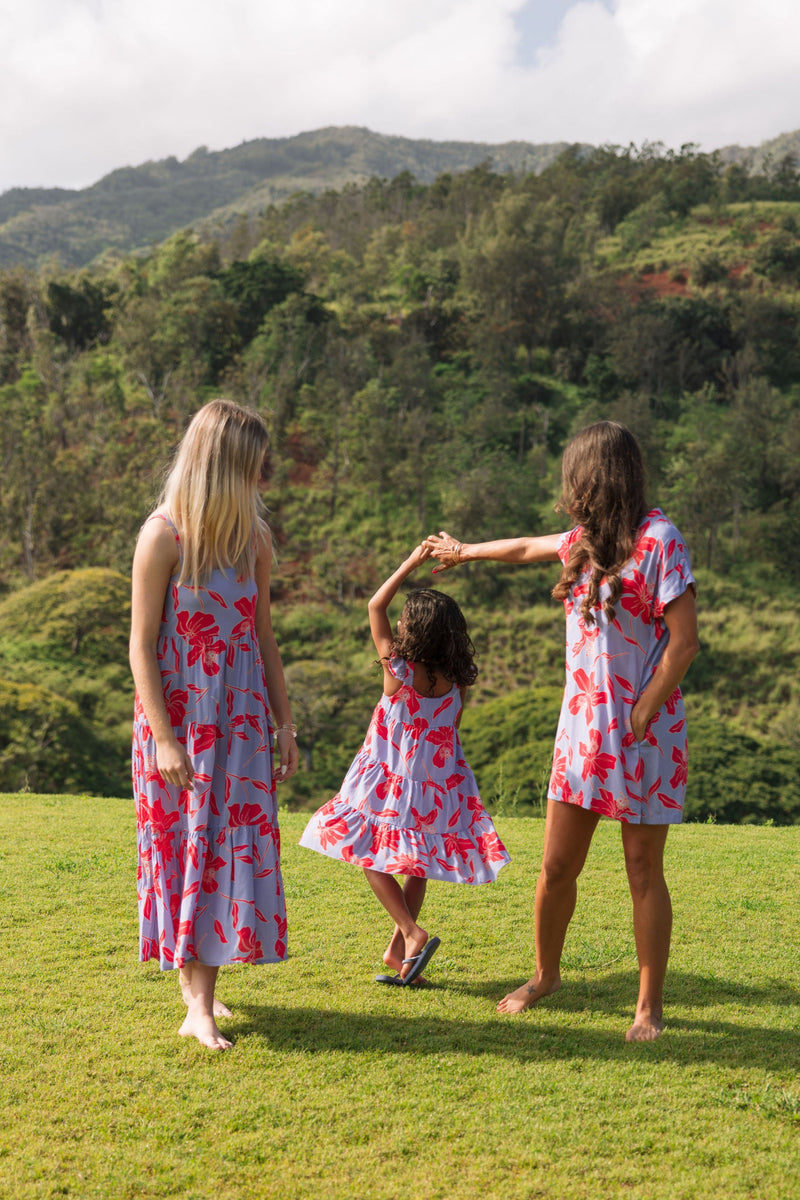 little girl with brown hair wearing a mid length dress with periwinkle background and bright red hibiscus print.