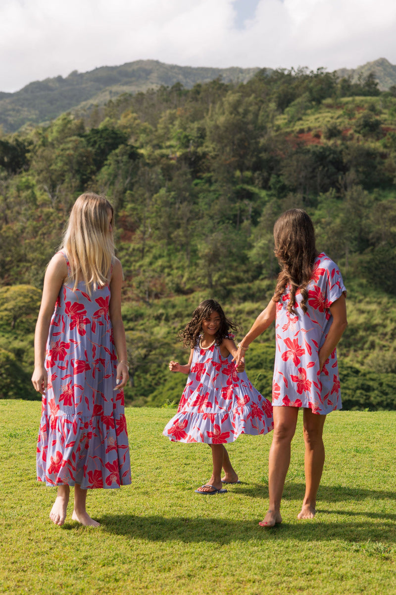 little girl with brown hair wearing a mid length dress with periwinkle background and bright red hibiscus print.