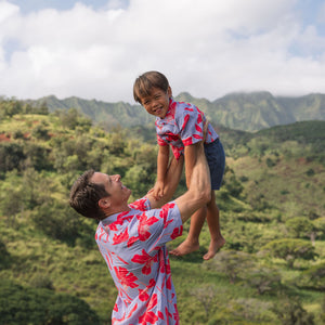 little boy with brown hair wearing a button up aloha shirt with periwinkle background and bright red hibiscus print.