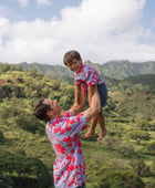 little boy with brown hair wearing a button up aloha shirt with periwinkle background and bright red hibiscus print.