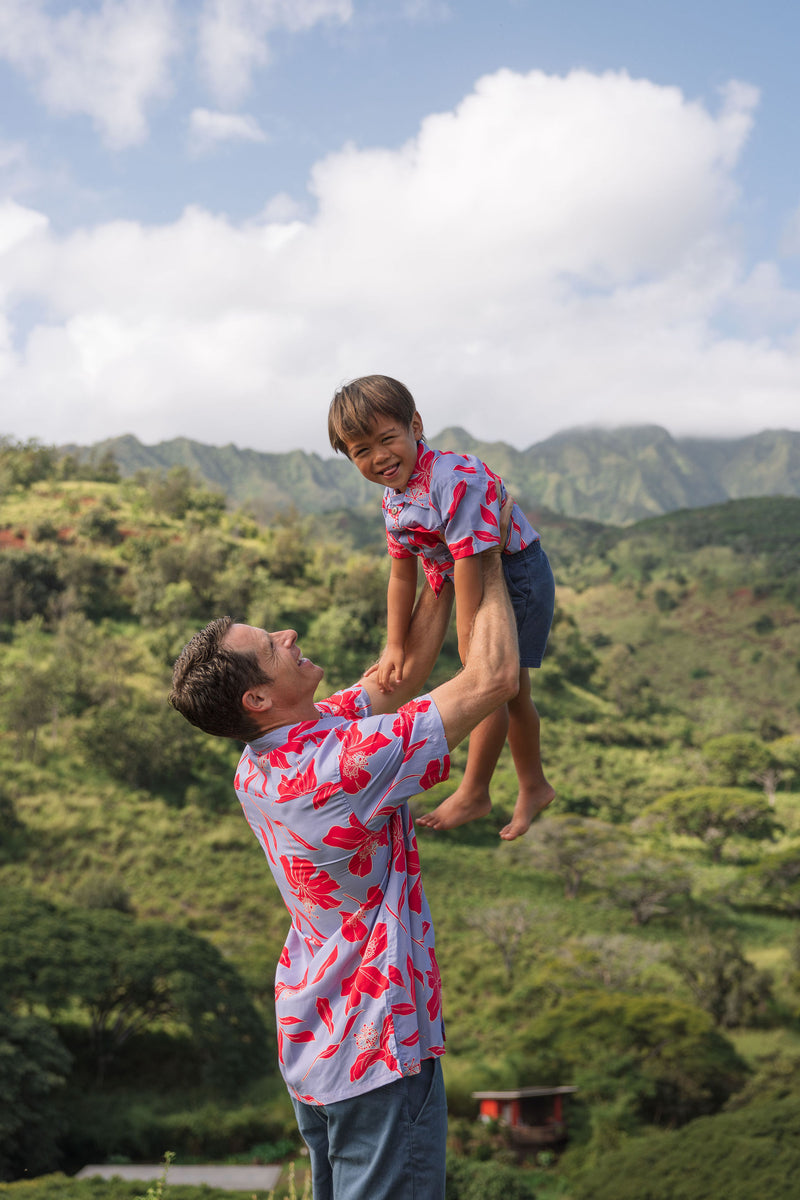 little boy with brown hair wearing a button up aloha shirt with periwinkle background and bright red hibiscus print.