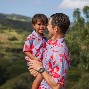 little boy with brown hair wearing a button up aloha shirt with periwinkle background and bright red hibiscus print.