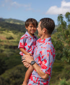 little boy with brown hair wearing a button up aloha shirt with periwinkle background and bright red hibiscus print.