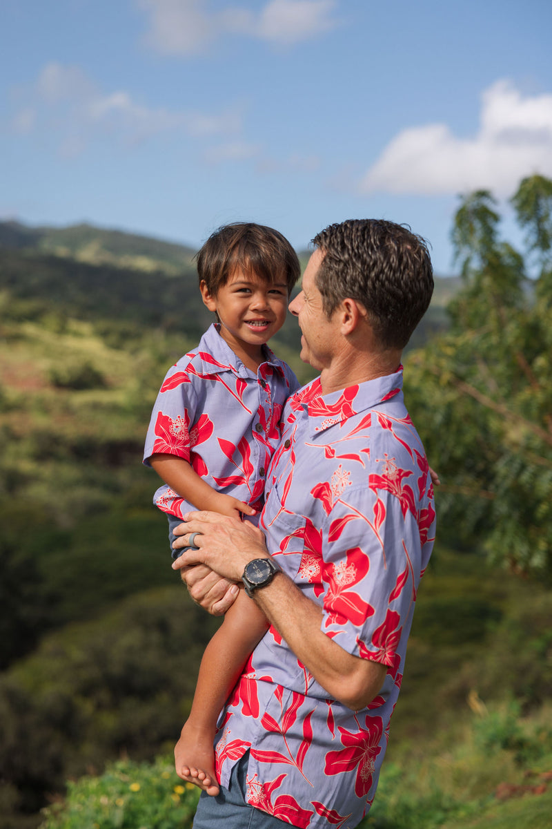 little boy with brown hair wearing a button up aloha shirt with periwinkle background and bright red hibiscus print.