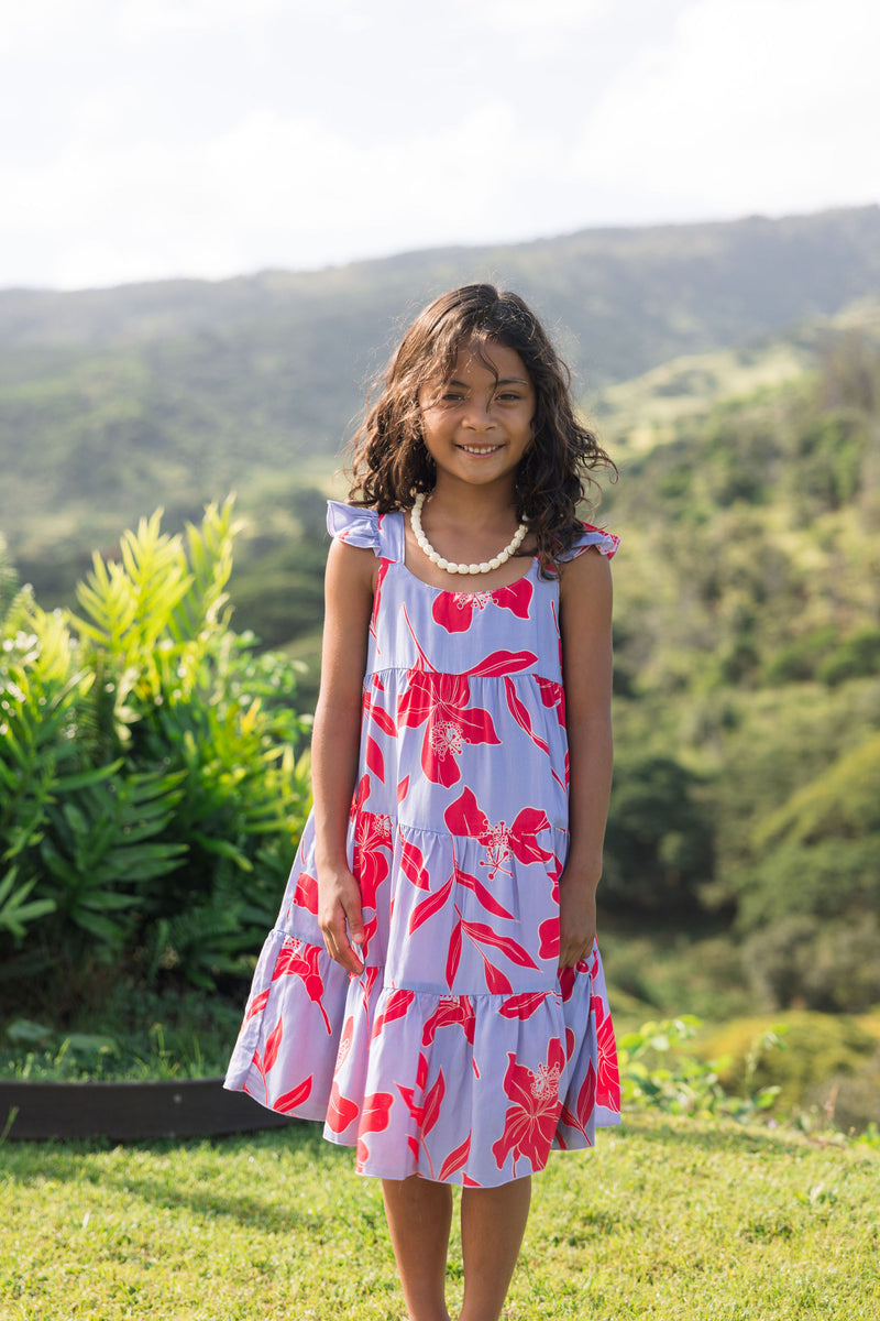 little girl with brown hair wearing a mid length dress with periwinkle background and bright red hibiscus print.