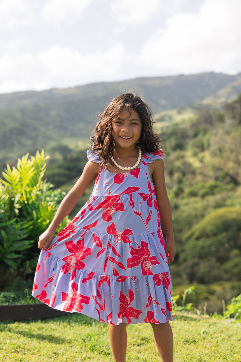 little girl with brown hair wearing a mid length dress with periwinkle background and bright red hibiscus print.