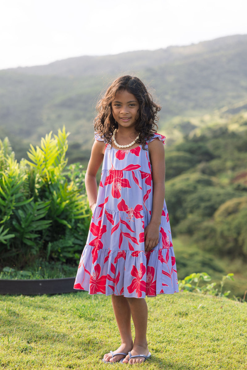 little girl with brown hair wearing a mid length dress with periwinkle background and bright red hibiscus print.