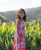 little girl with brown hair wearing a mid length dress with periwinkle background and bright red hibiscus print.