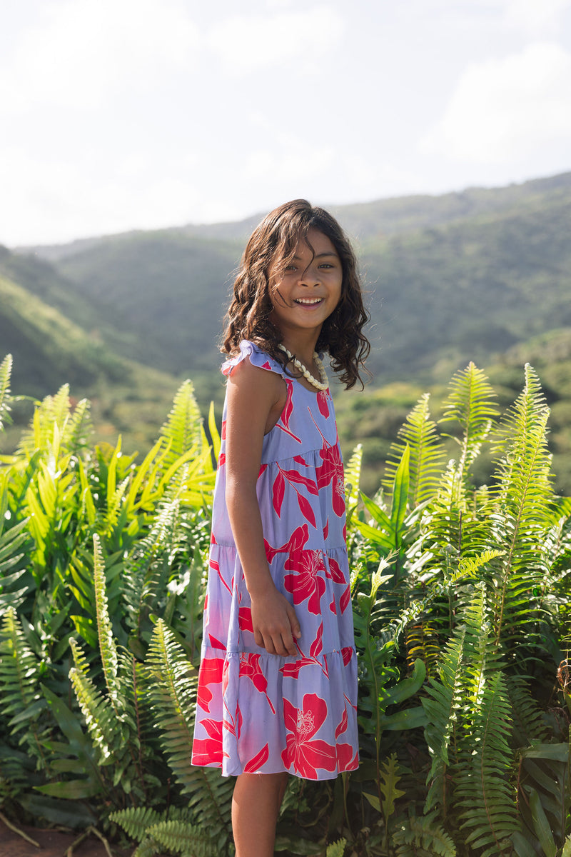 little girl with brown hair wearing a mid length dress with periwinkle background and bright red hibiscus print.