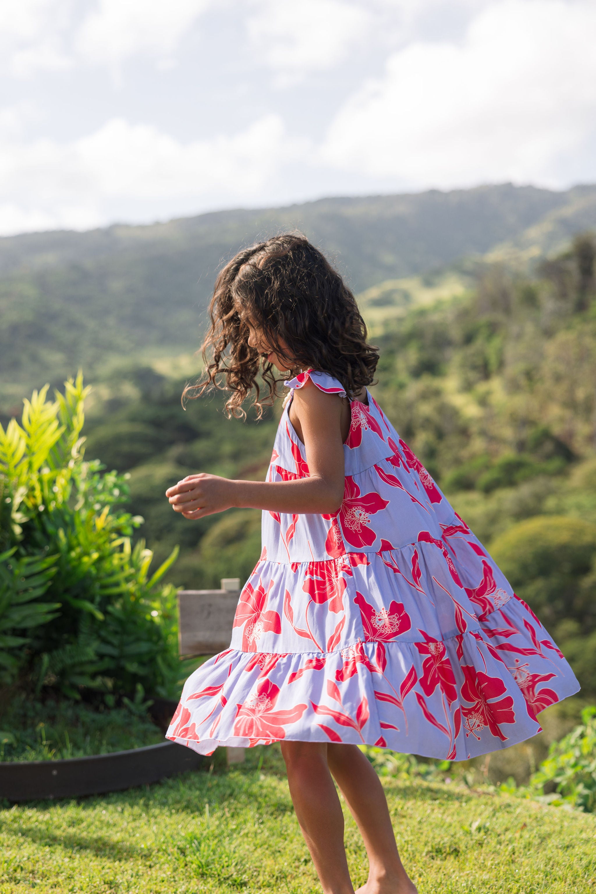 little girl with brown hair wearing a mid length dress with periwinkle background and bright red hibiscus print.
