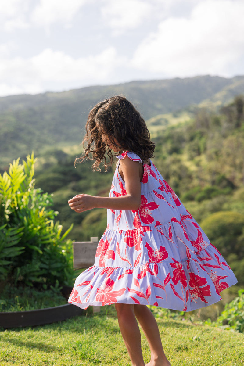 little girl with brown hair wearing a mid length dress with periwinkle background and bright red hibiscus print.