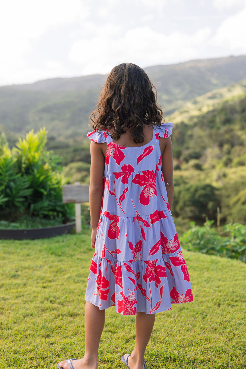 little girl with brown hair wearing a mid length dress with periwinkle background and bright red hibiscus print.