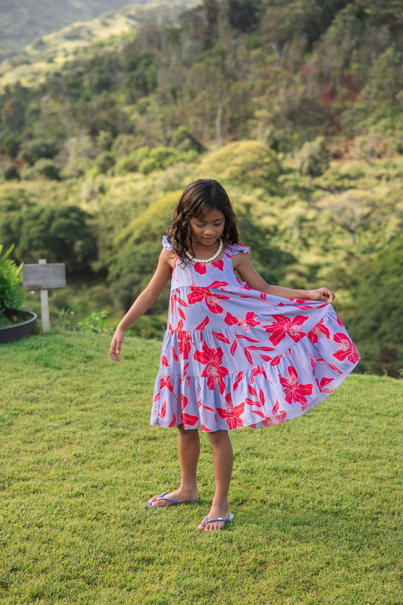 little girl with brown hair wearing a mid length dress with periwinkle background and bright red hibiscus print.