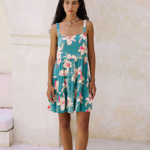 woman with brown hair wearing a tiered mini dress in teal with large cream colored orchids.