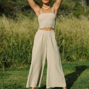 Woman wearing smocked tube top with spaghetti straps and loose pant in beige linen.