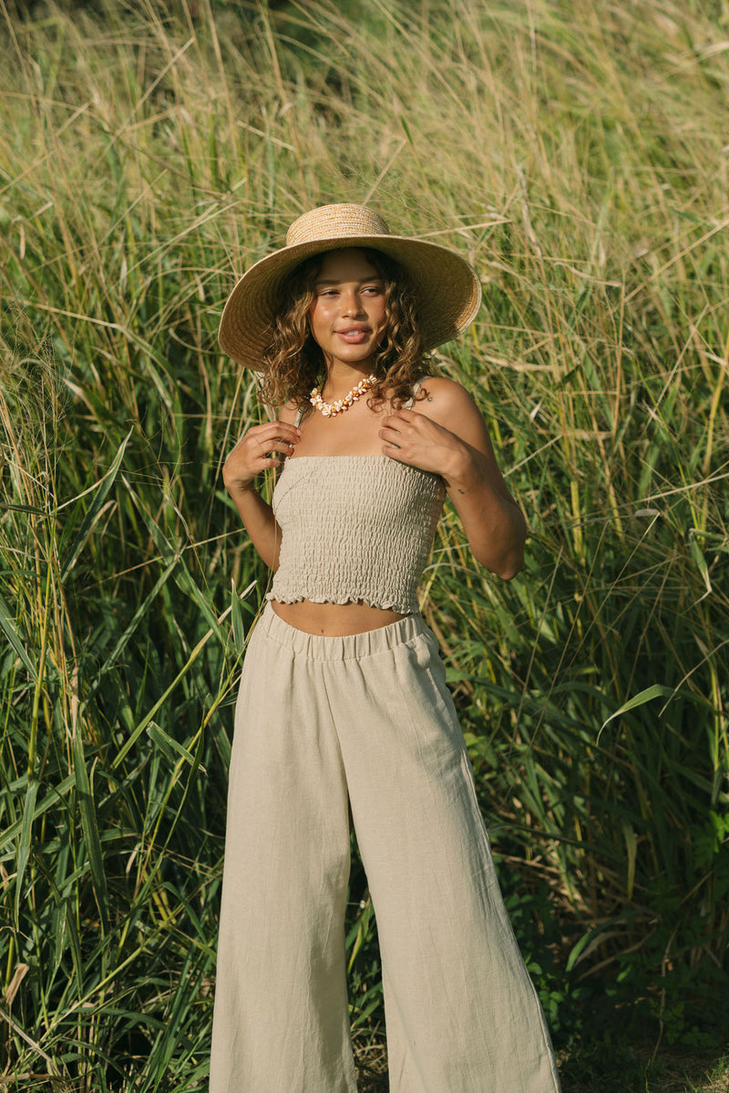 Woman wearing smocked tube top with spaghetti straps and loose pant in beige linen.