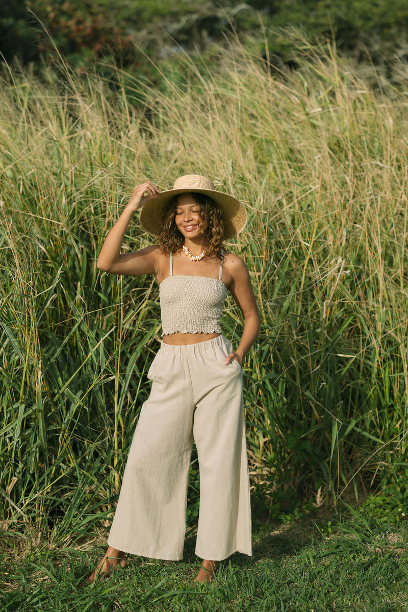 Woman wearing smocked tube top with spaghetti straps and loose pant in beige linen.