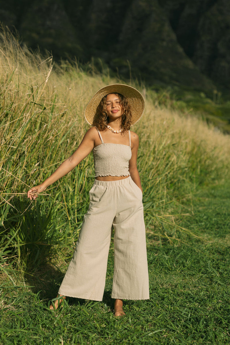 Woman wearing smocked tube top with spaghetti straps and loose pant in beige linen.