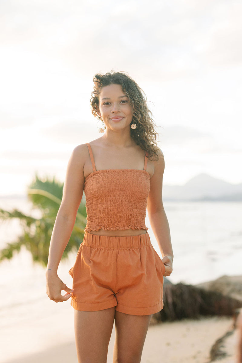 Woman wearing burnt orange linen smocked tube top with spaghetti straps and short.