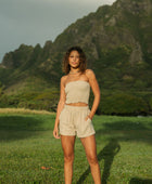 Woman wearing smocked beige linen tube top with spaghetti straps and shorts.