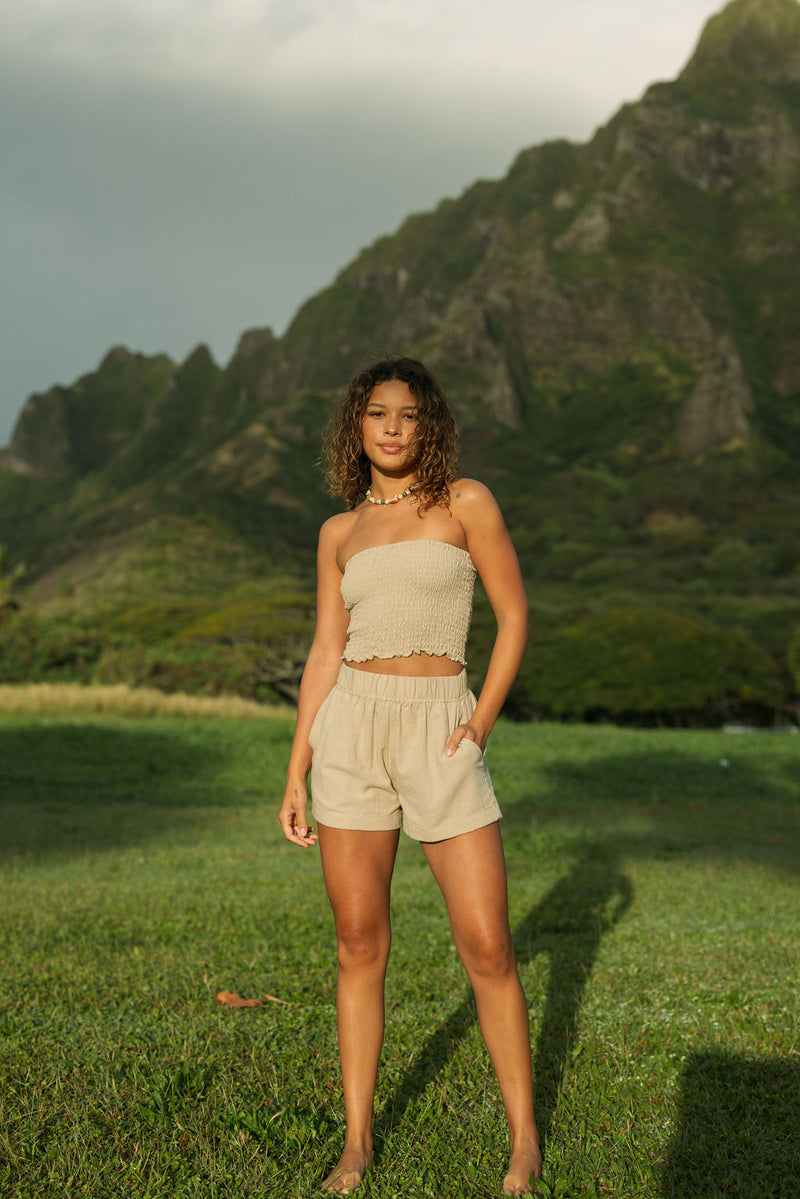 Woman wearing smocked beige linen tube top with spaghetti straps and shorts.