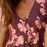 woman with brown hair wearing brown/maroon toned button up romper with pink lei awapuhi print