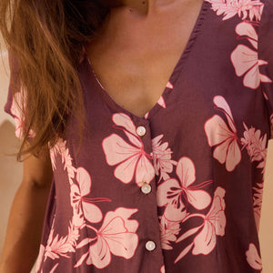 woman with brown hair wearing brown/maroon toned button up romper with pink lei awapuhi print