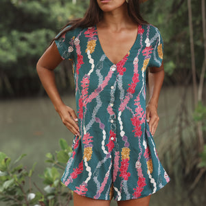 woman with dark brown hair wearing button up romper in dark teal with shell and lei print