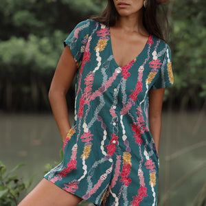 woman with dark brown hair wearing button up romper in dark teal with shell and lei print