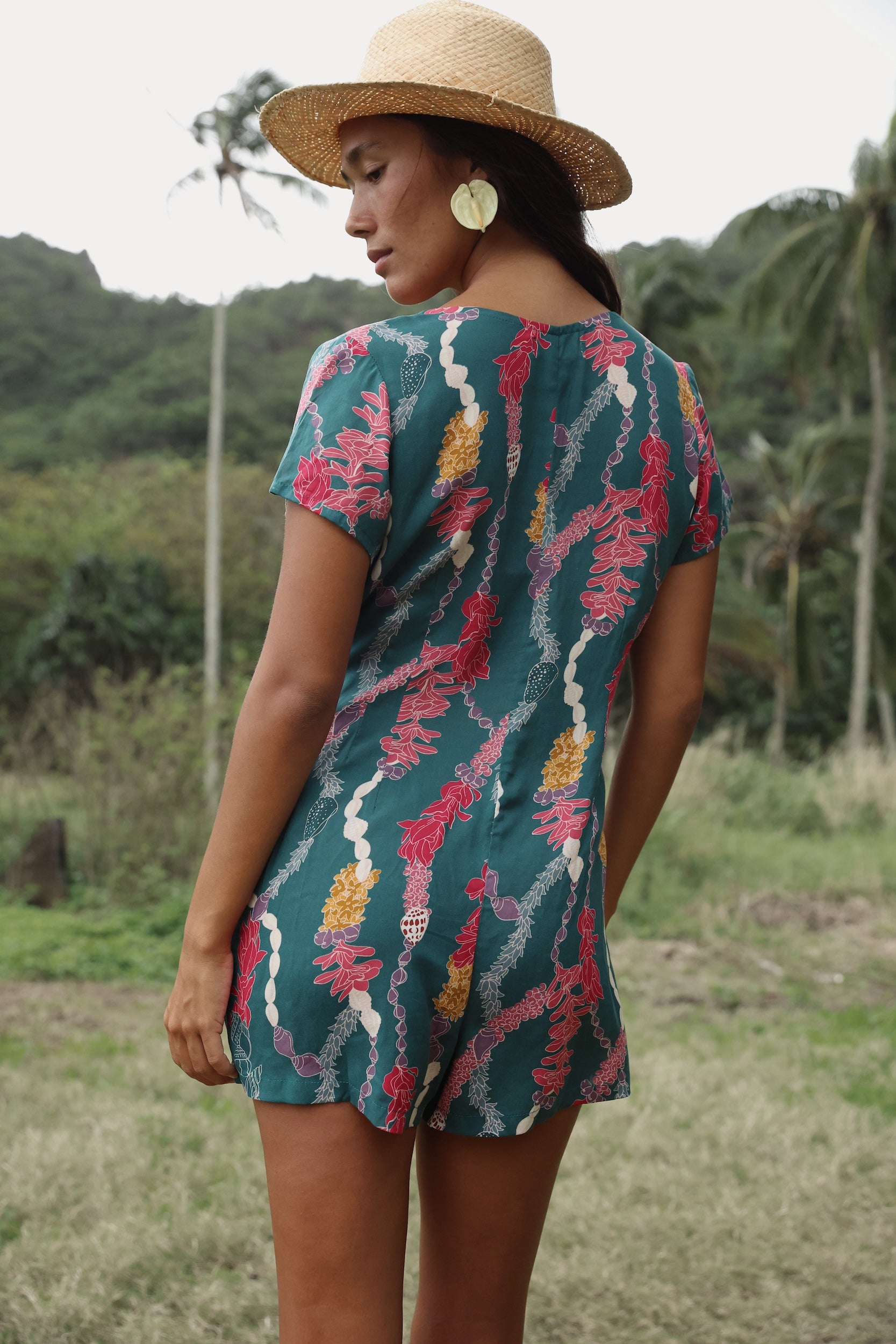 woman with dark brown hair wearing button up romper in dark teal with shell and lei print