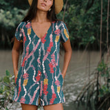 woman with dark brown hair wearing button up romper in dark teal with shell and lei print