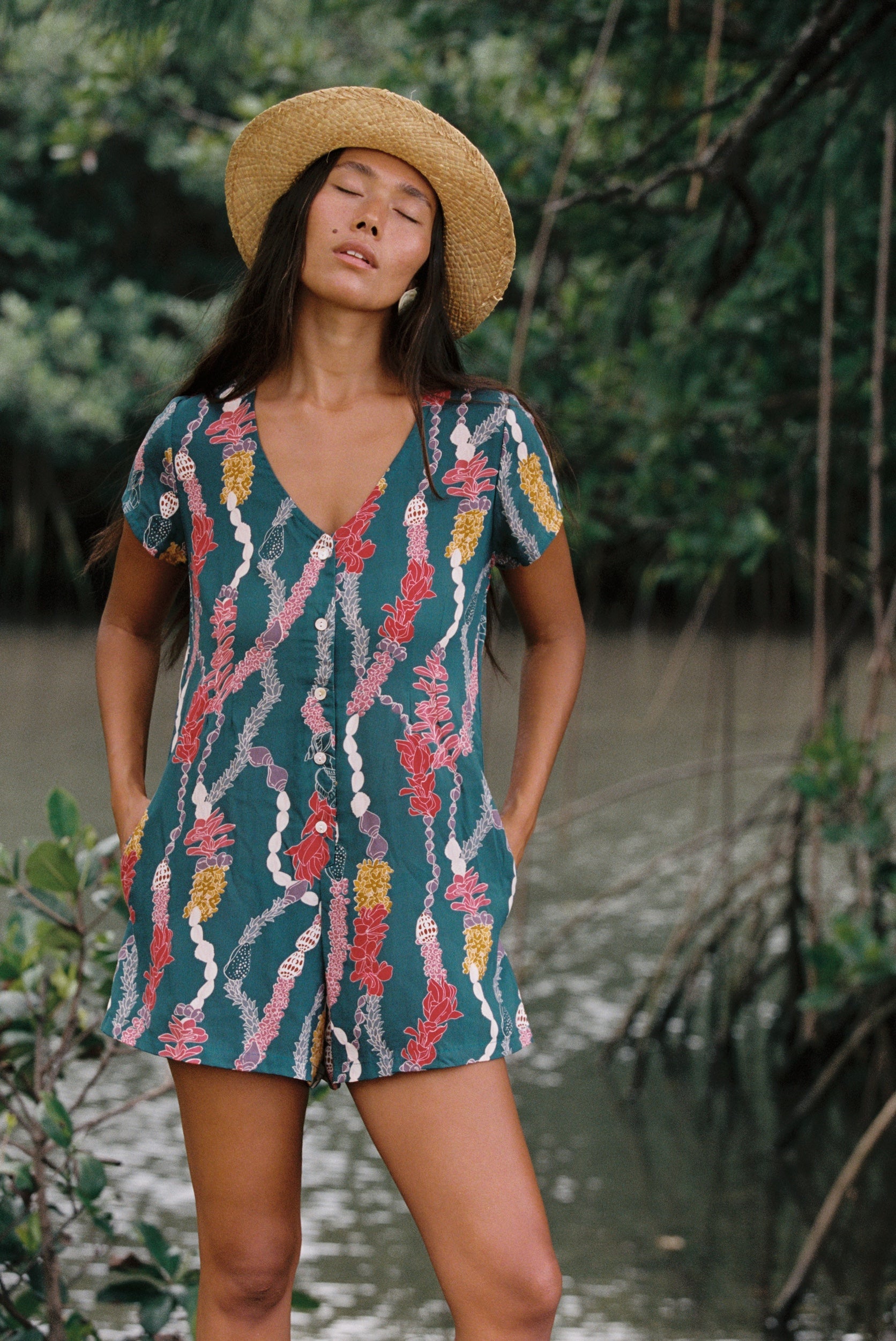 woman with dark brown hair wearing button up romper in dark teal with shell and lei print