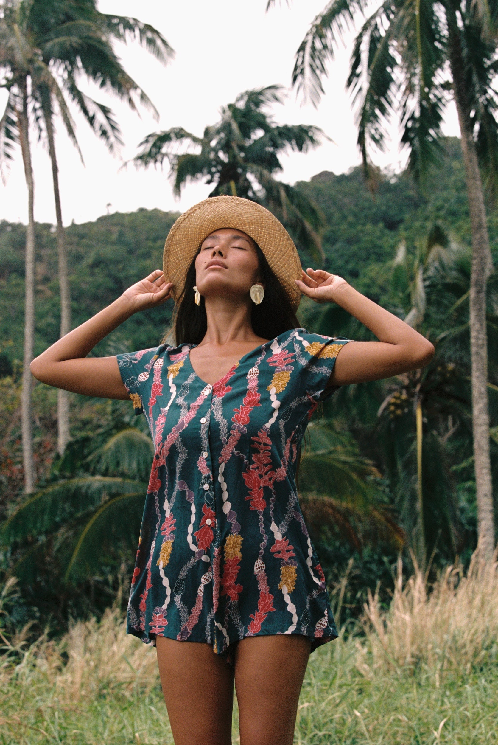 woman with dark brown hair wearing button up romper in dark teal with shell and lei print