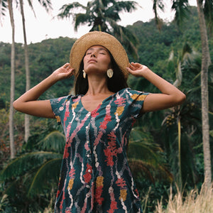 woman with dark brown hair wearing button up romper in dark teal with shell and lei print