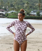 Woman with brown hair wearing a high neck long sleeve one piece bikini in blush and brown hibiscus print.