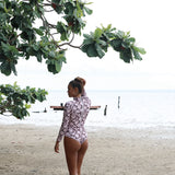 Woman with brown hair wearing a high neck long sleeve one piece bikini in blush and brown hibiscus print.