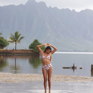 Woman with brown hair wearing a bikini top with a knot detail and a high waisted bikini bottoms in blush and brown hibiscus print.