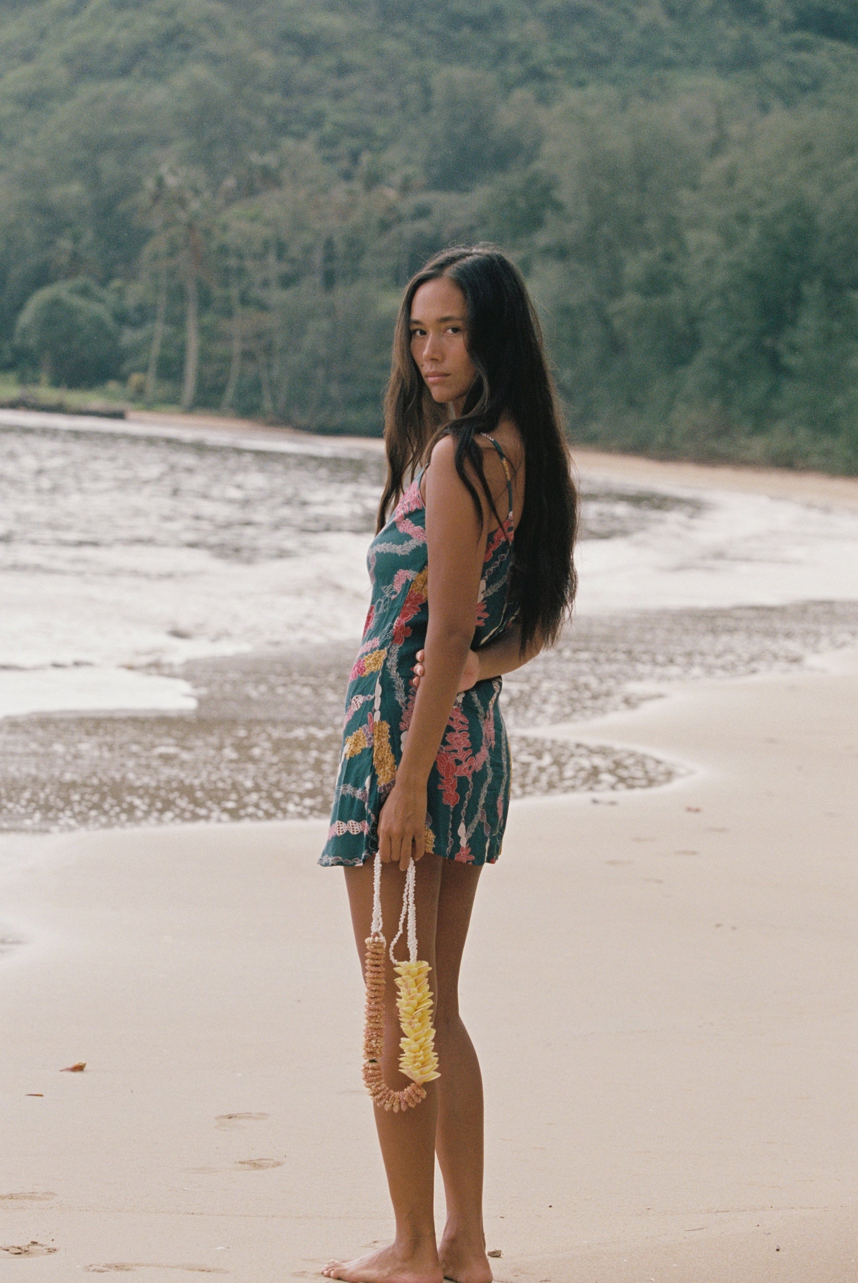 woman with dark brown hair wearing mini spaghetti strap dress in dark teal with shell and lei print