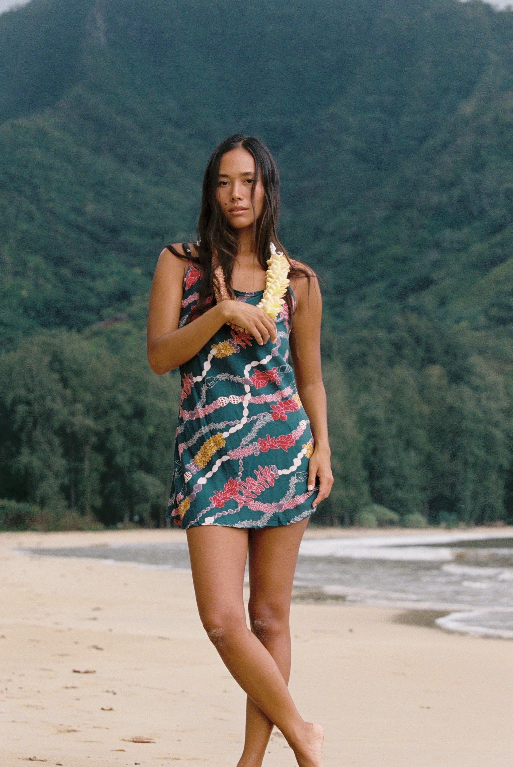 woman with dark brown hair wearing mini spaghetti strap dress in dark teal with shell and lei print