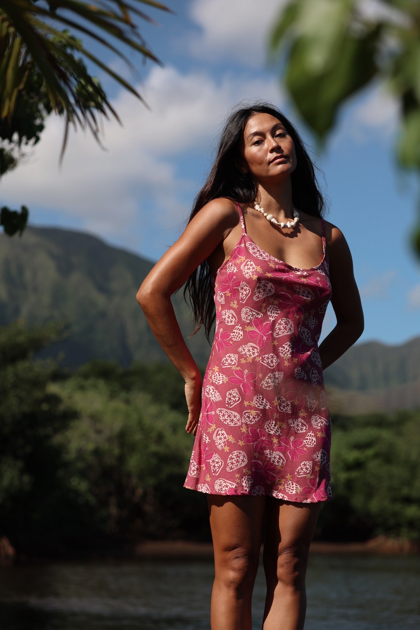 woman with brown hair wearing a pink spaghetti strap scoop neck mini dress with tie back detail in a shell and pua print 