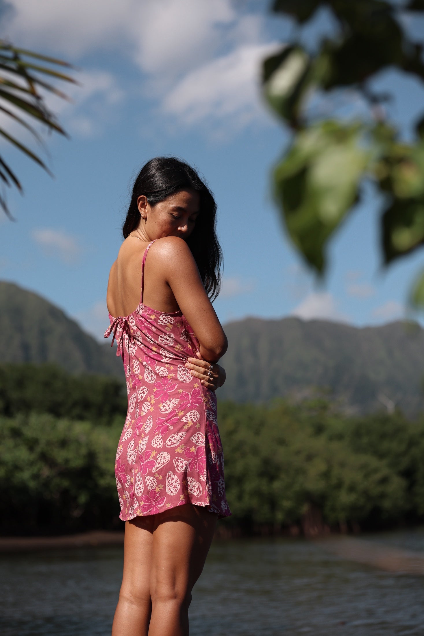 woman with brown hair wearing a pink spaghetti strap scoop neck mini dress with tie back detail in a shell and pua print 