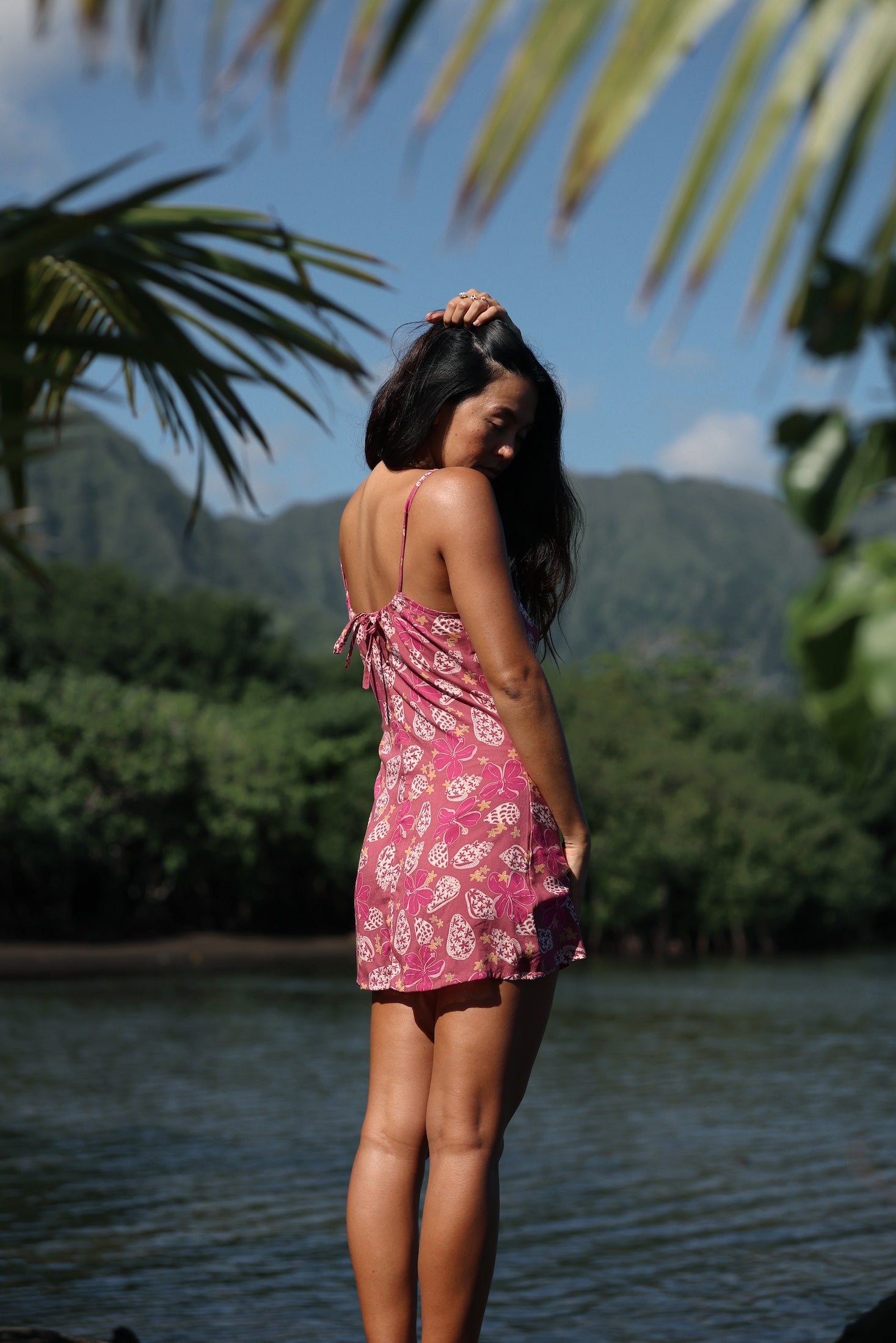 woman with brown hair wearing a pink spaghetti strap scoop neck mini dress with tie back detail in a shell and pua print 