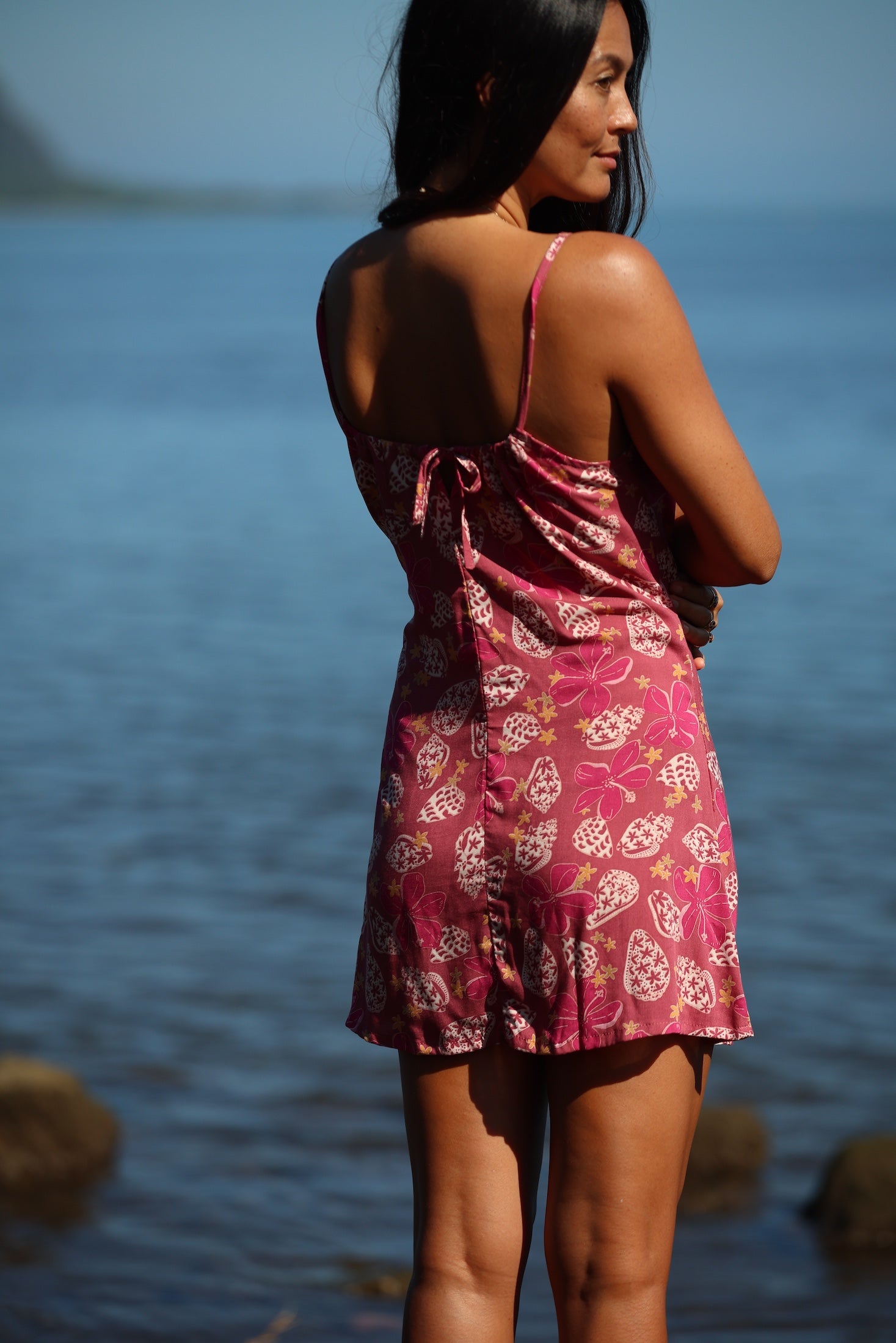 woman with brown hair wearing a pink spaghetti strap scoop neck mini dress with tie back detail in a shell and pua print 