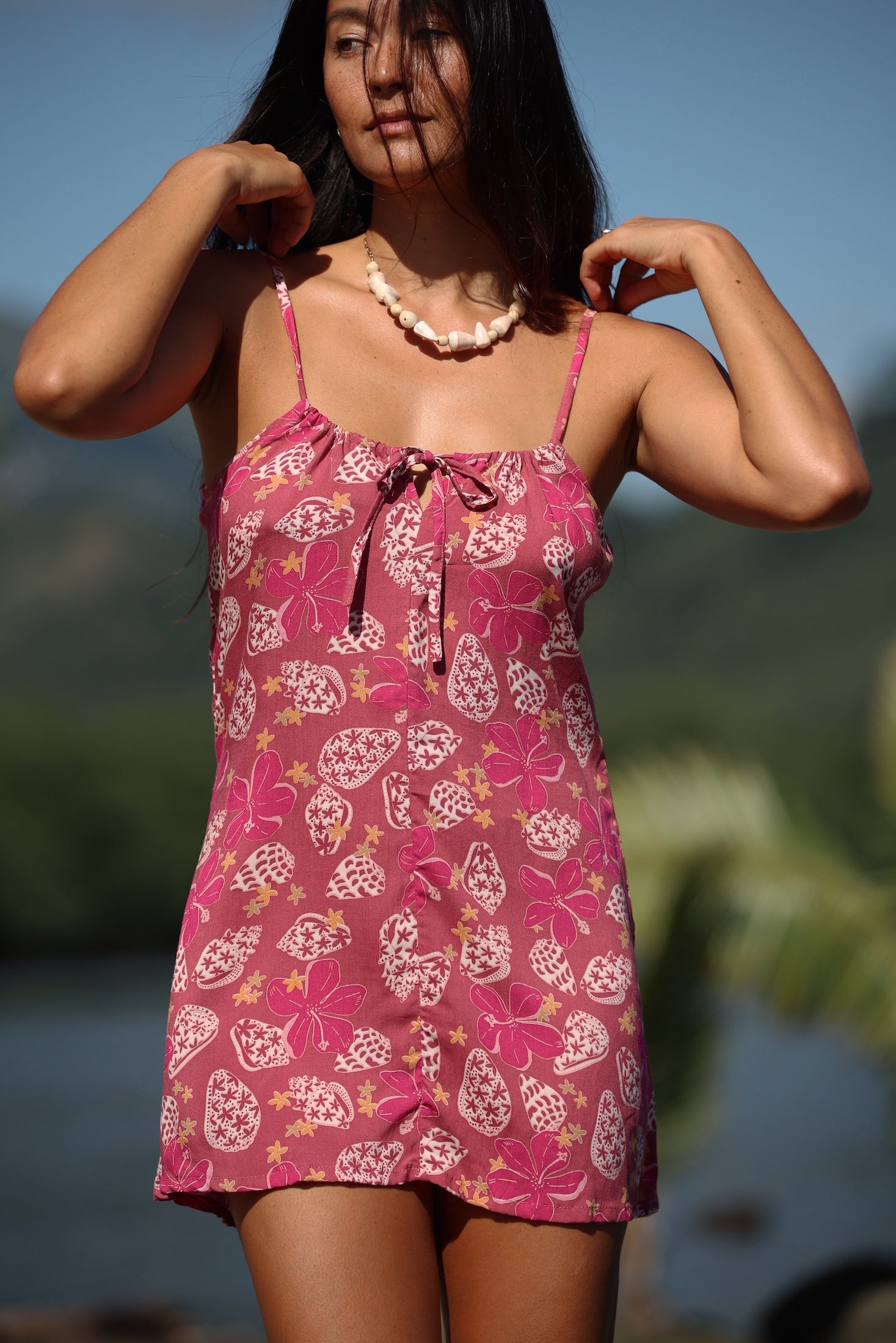 woman with brown hair wearing a pink spaghetti strap scoop neck mini dress with tie back detail in a shell and pua print 